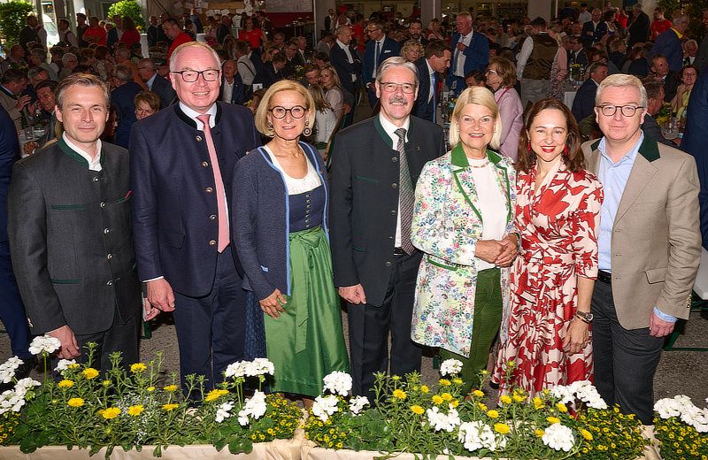 Sechs Personen stehen bei einem formellen Event mit Blumen vor ihnen. Dahinter versammelt sich eine Menschenmenge in einem großen Saal.