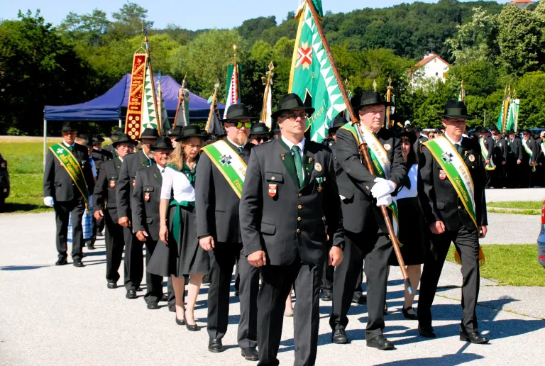 A marching band in formal attire, carrying flags and sashes, parades on a road with trees and buildings in the background.