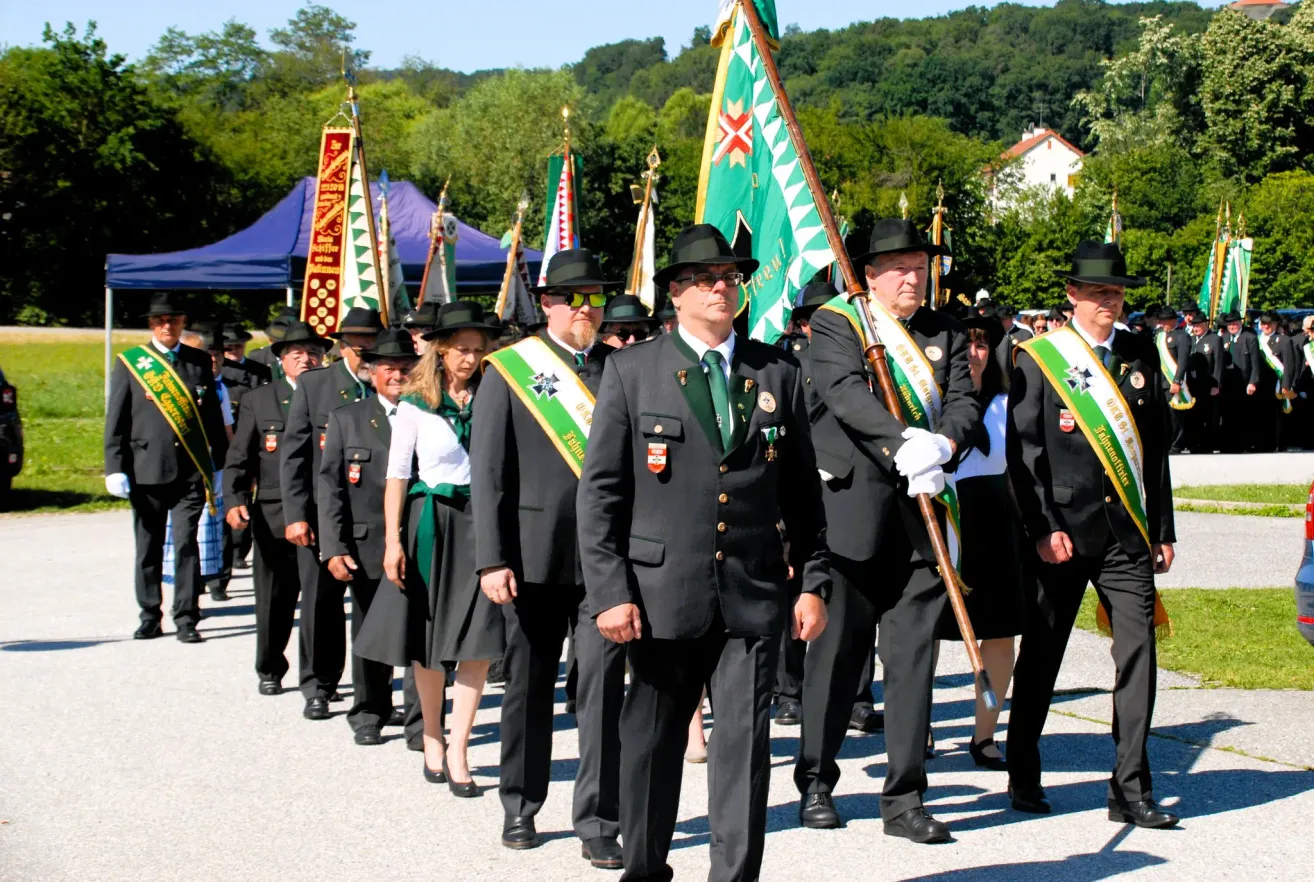 A marching band in formal attire, carrying flags and sashes, parades on a road with trees and buildings in the background.