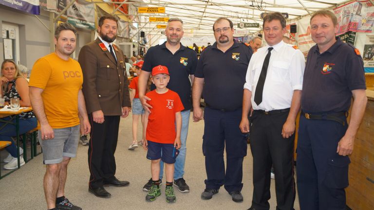 Fünf Männer in Uniformen stehen in einem Korridor, mit einem kleinen Jungen in einem orangefarbenen Shirt zwischen zwei Männern. Sie alle scheinen für ein Foto zu posieren.