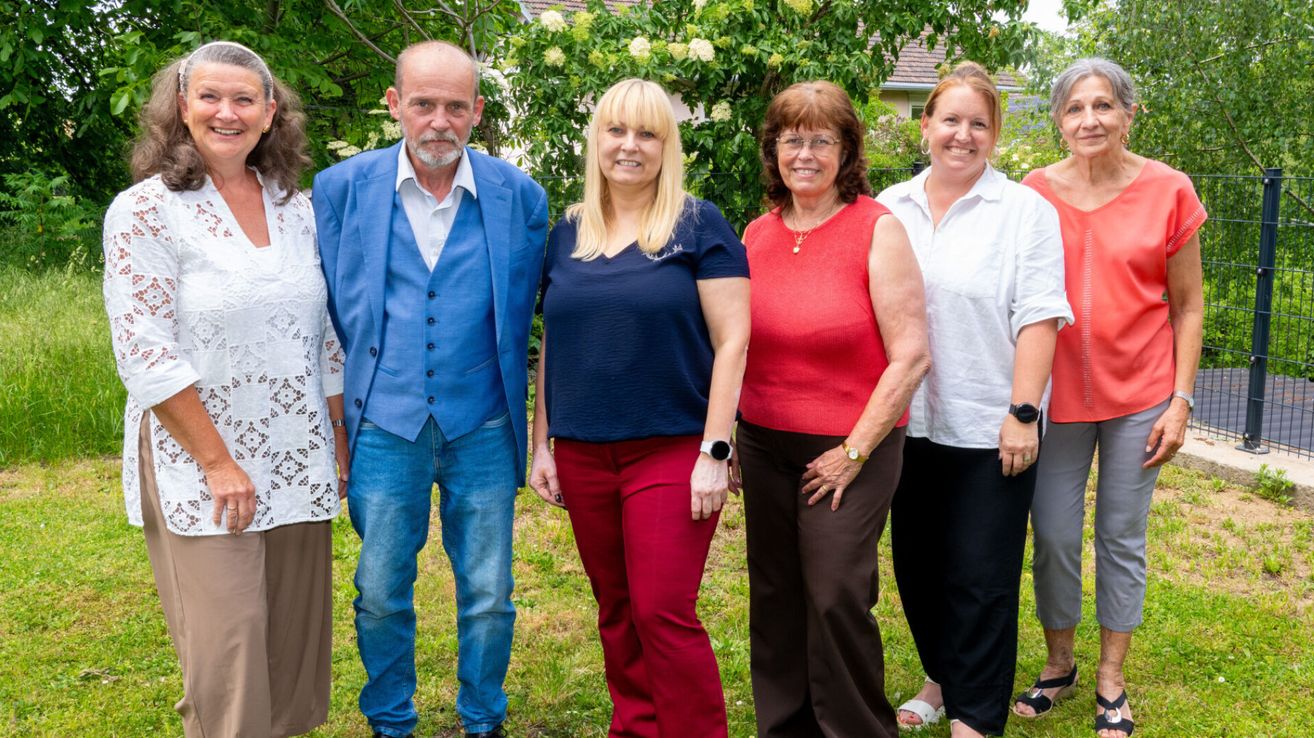 Five people pose for a photo in a garden, smiling. A man in blue and a woman in red stand in the middle. Trees and greenery are in the background.