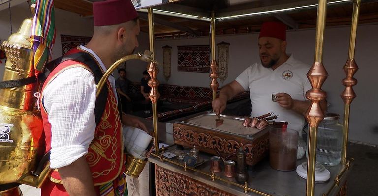 Zwei Männer bereiten Getränke in einem Marktstand zu. Einer trägt einen roten Fez und hält eine Kanne, der andere in einem weißen Hemd hält einen Becher.