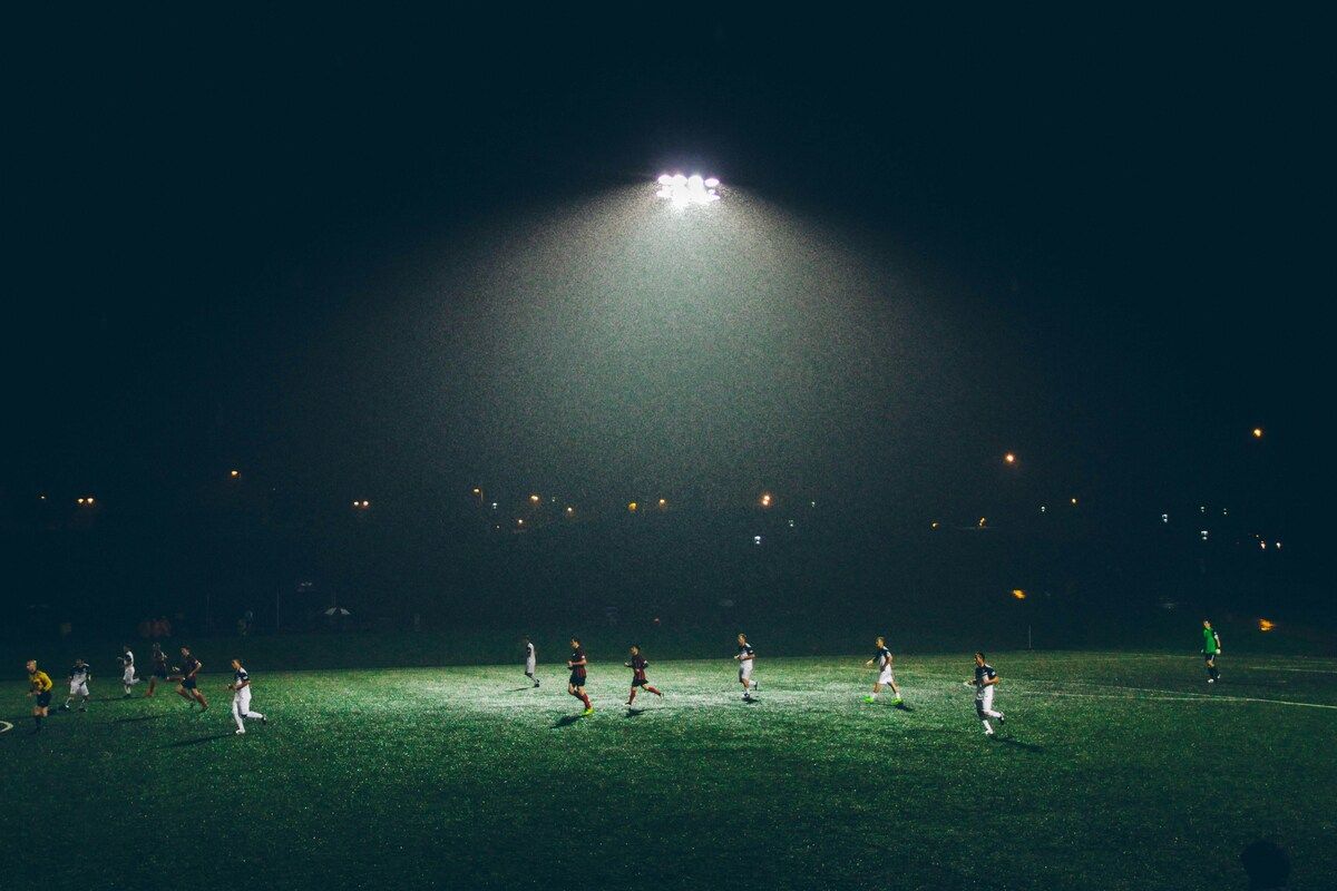 Ein Nachtfußballspiel mit Spielern, die auf einem Feld unter hellen Lichtern und einem regnerischen Himmel laufen.