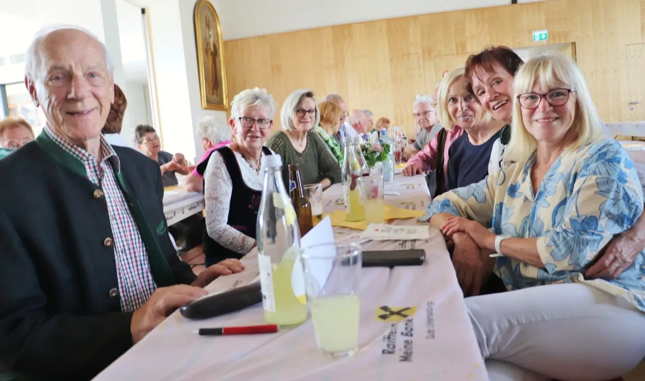 Eine Gruppe älterer Menschen sitzt an einem Tisch mit Getränken und Blumen. Sie lächeln und posieren für ein Foto.