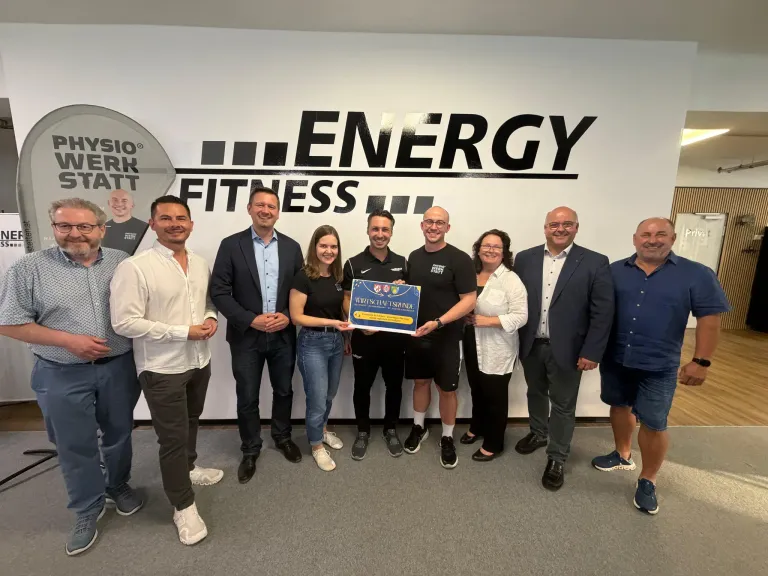 A group of people pose for a photo in front of a wall with the Energy Fitness logo, holding a certificate.