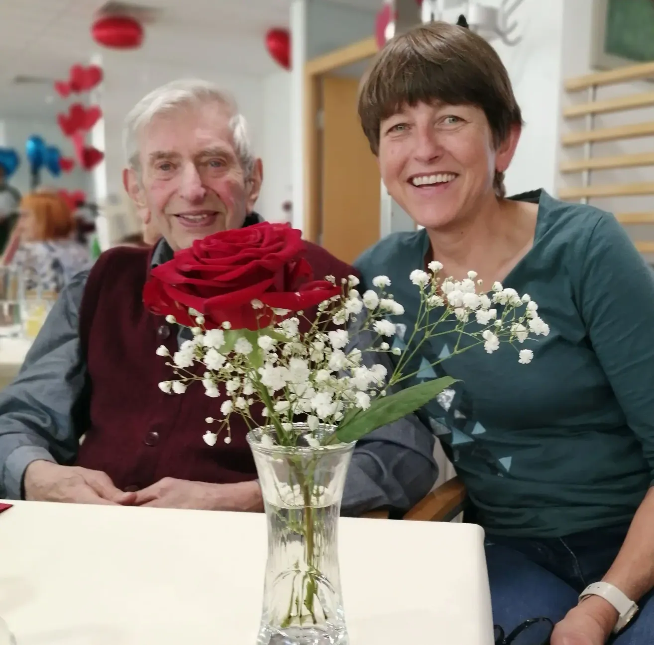 An elderly man and woman are smiling and posing for a photo with a vase of red roses and baby's breath. They are seated at a table. Behind them, there are people and red balloons hanging from the ceiling.