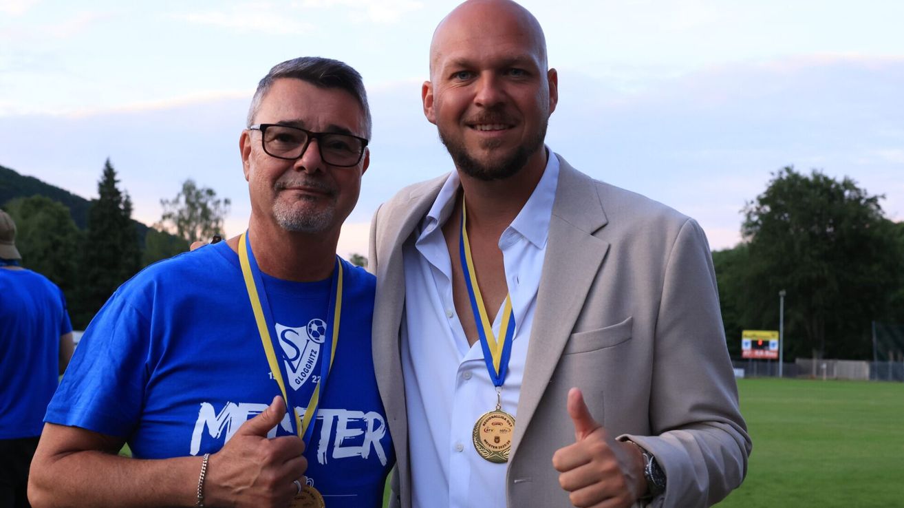 Two men stand next to each other at a sports event. One wears a blue shirt and medals, while the other wears a suit and gives a thumbs up.