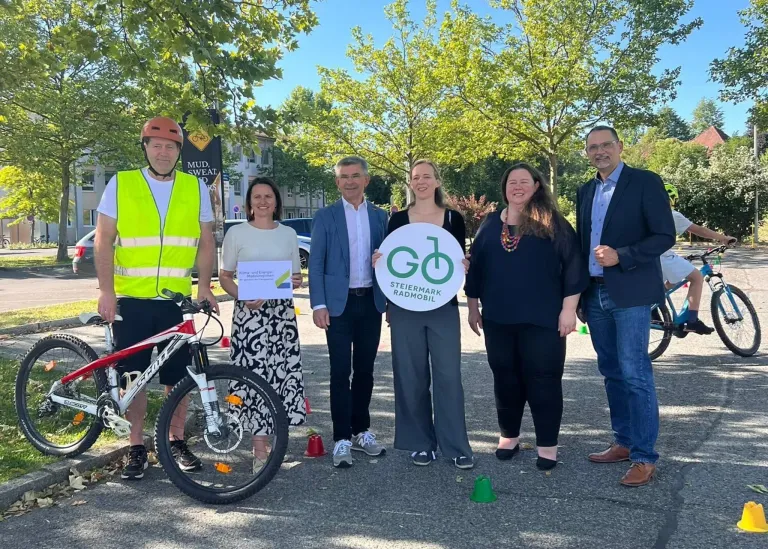 A group of people in casual attire stand in a parking lot, holding a green circular sign with GD and Steiermark Radmobil. One holds a paper, and others have bicycles. They stand next to cones.