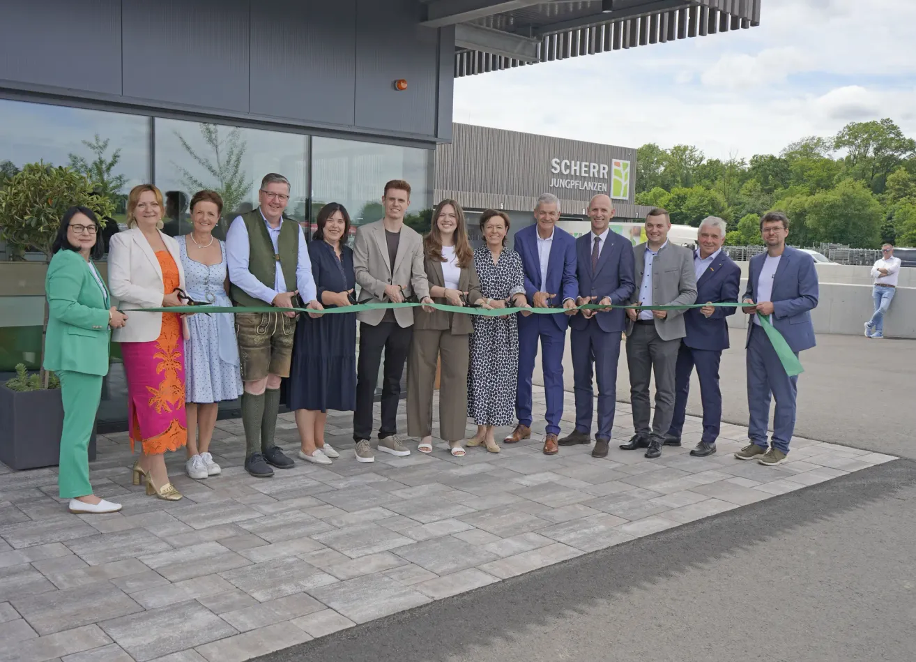 A group of people, possibly business associates, pose in front of a building, holding a green ribbon for a ribbon-cutting ceremony.
