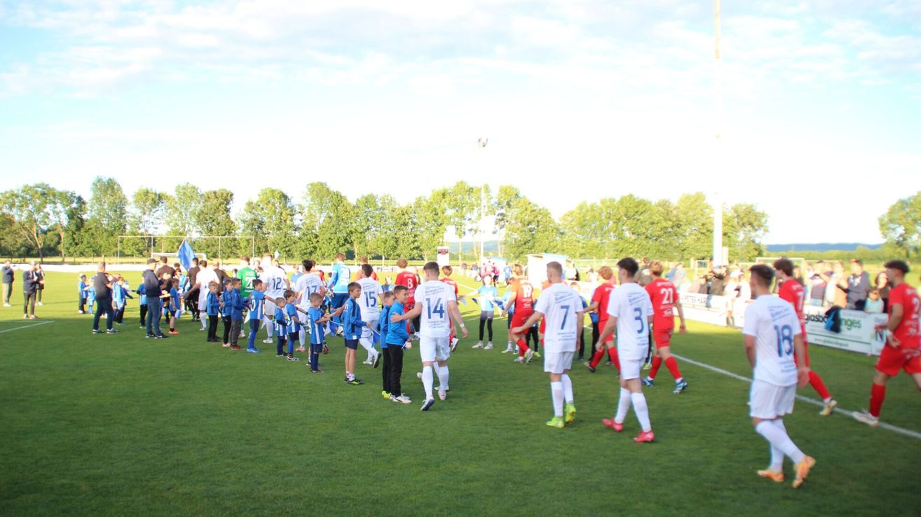 Eine Gruppe von Fußballspielern, sowohl Erwachsene als auch Kinder, tragen Trikots mit Nummern und stehen auf einem Rasenfeld. Die Spieler stehen in einer Reihe, wahrscheinlich für ein Teamfoto. Das Feld ist von Bäumen umgeben.