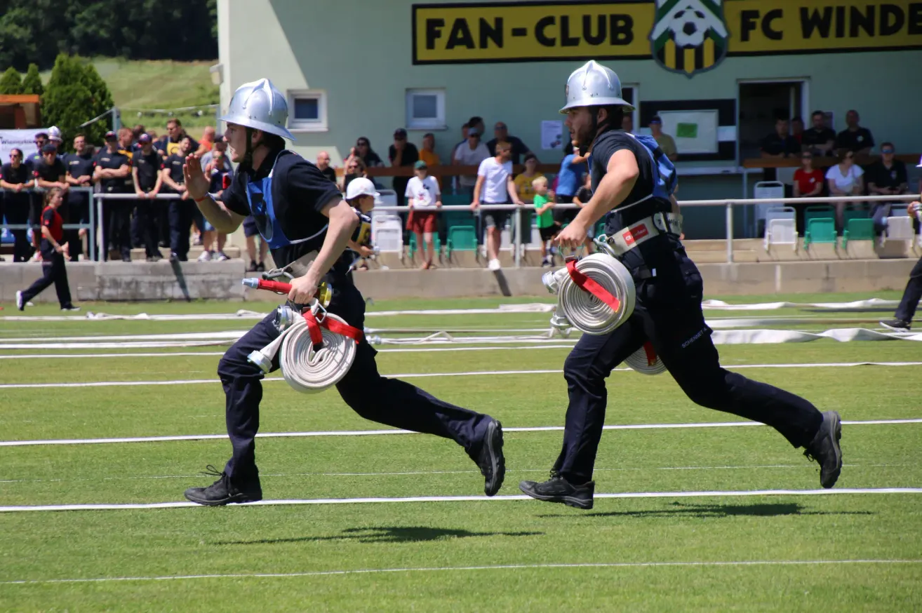 Two firefighters in uniform and helmets run across a field with a fire hose, with a crowd watching.