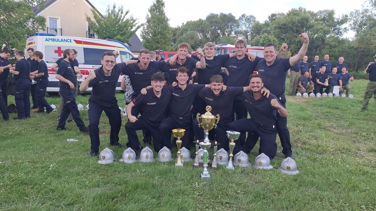 Eine Gruppe von Männern in Uniform, möglicherweise Feuerwehrleute, posiert mit ihren Trophäen für ein Foto. Sie befinden sich auf einem Grasfeld mit Helmen, Feuerwehrwagen und Bäumen im Hintergrund.