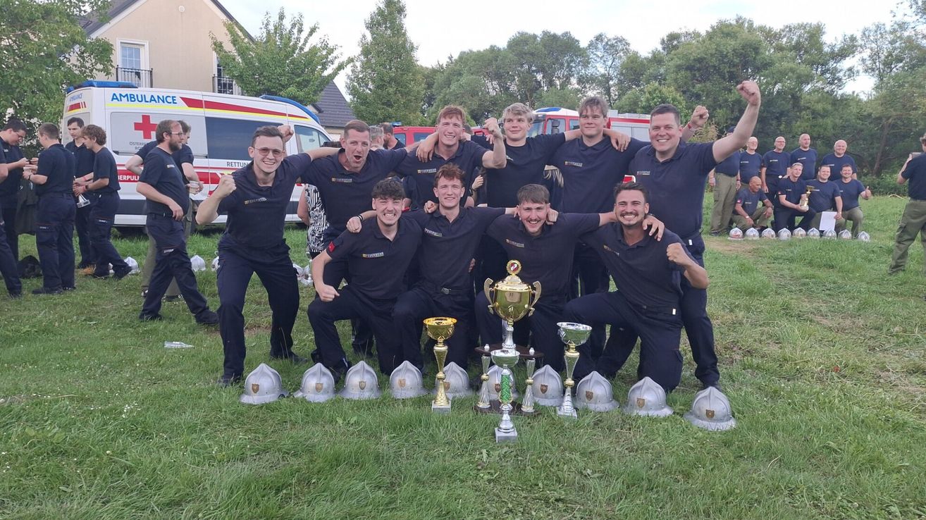 Eine Gruppe von Männern in Uniform, möglicherweise Feuerwehrleute, posiert mit ihren Trophäen für ein Foto. Sie befinden sich auf einem Grasfeld mit Helmen, Feuerwehrwagen und Bäumen im Hintergrund.