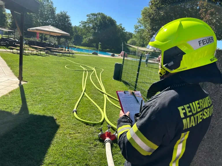 Ein Feuerwehrmann inspiziert den Feuerhydranten mit einem Klemmbrett. Mehrere Schläuche liegen auf dem Rasen. Dahinter befindet sich ein Schwimmbad mit Zaun.