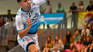 Ein männlicher Athlet in einem weißen und blauen Trikot springt in die Luft und wirft einen Ball während eines Spiels. Zuschauer sitzen hinter ihm und schauen sich das Spiel an.