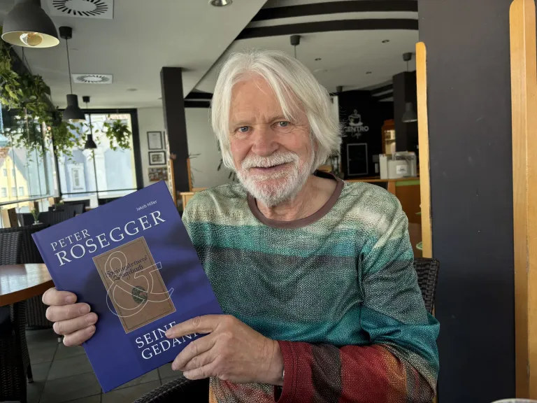 An elderly man in a striped sweater holds a blue book titled 'Seine Gedanken' and smiles for the camera in an indoor setting.