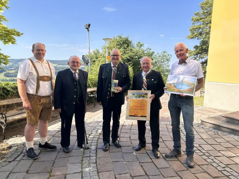 Fünf Männer in formeller Kleidung stehen auf einem gepflasterten Weg und lächeln. Einer hält einen gerahmten Preis, ein anderer eine Malerei. Einer trägt traditionelle bayerische Kleidung. Bäume und ein klarer Himmel im Hintergrund.