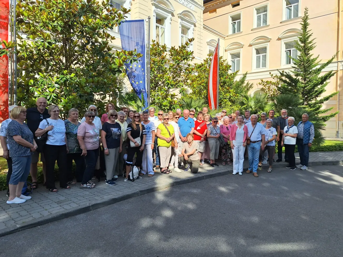 Eine Gruppe von Menschen steht vor einem Gebäude mit Flaggen, Bäumen und Pflanzen an einem sonnigen Tag. Sie tragen Freizeitkleidung und scheinen für ein Gruppenfoto zu posieren.