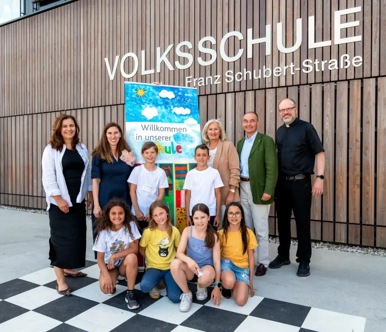 A group of people stand outside a building with 'Volksschule Franz Schubert-Straße' on it. They are smiling and posing for a photo.