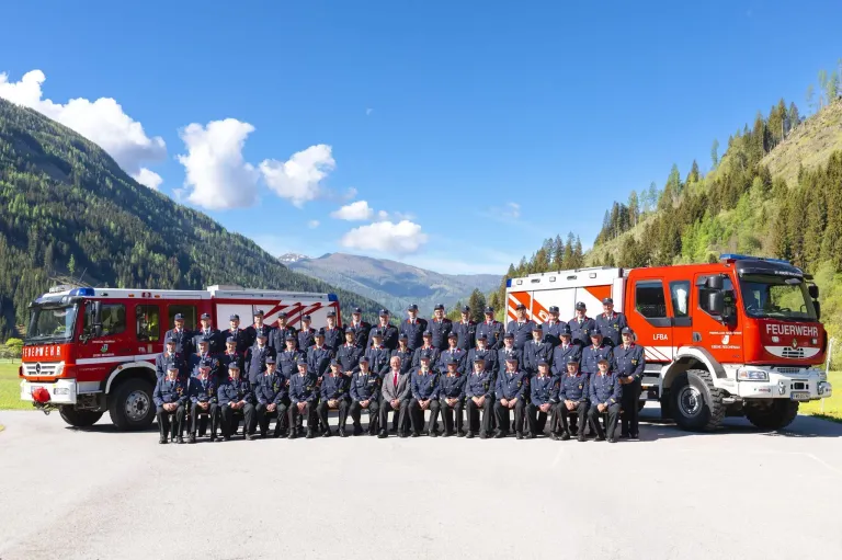 A group of firefighters in uniform are posing for a photo in front of two fire trucks with mountains in the background.