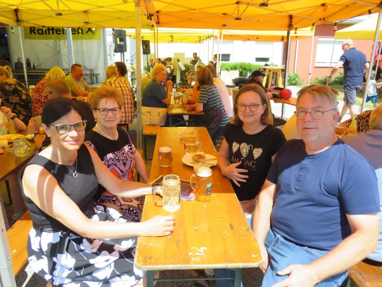Three women and a man sit at a table under a yellow tent, surrounded by other people at an outdoor event. They have drinks and plates in front of them.
