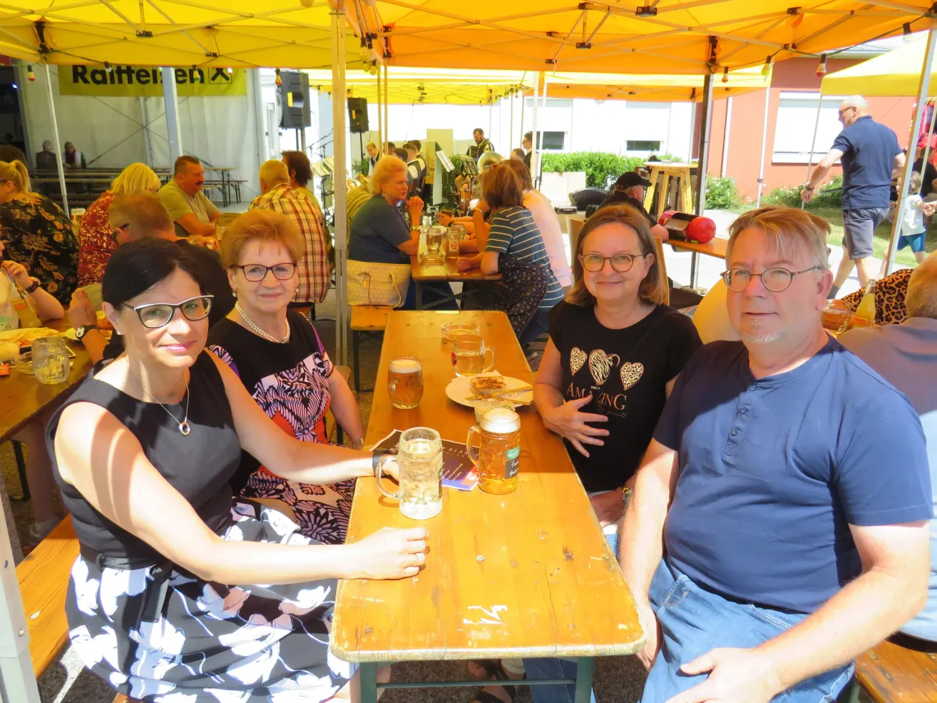 Three women and a man sit at a table under a yellow tent, surrounded by other people at an outdoor event. They have drinks and plates in front of them.