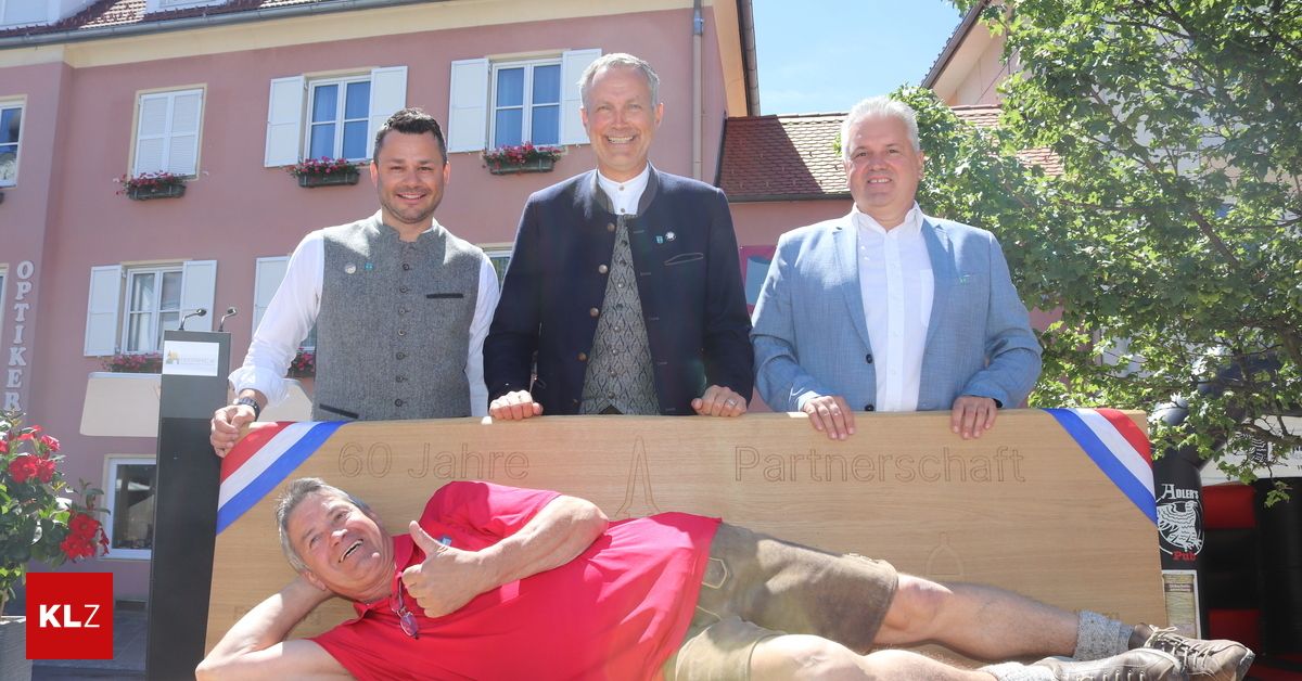 Four men stand behind a wooden sign saying 60 Jahre Partnerschaft. They all wear smiles. The man lying down gives a thumbs up.