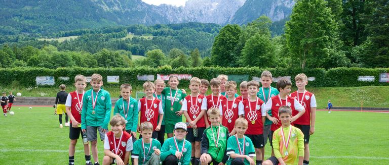 A group of young boys wearing medals pose for a picture on a grassy field with mountains in the background.