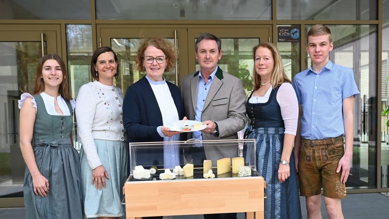 A group of people are smiling and posing for a photo in front of a table with various cheeses in a glass case. A man holds a plate with cheese, and a woman wears a necklace. Behind them are glass doors and a signboard.