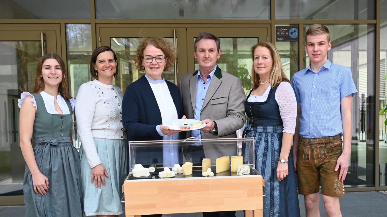 A group of people are smiling and posing for a photo in front of a table with various cheeses in a glass case. A man holds a plate with cheese, and a woman wears a necklace. Behind them are glass doors and a signboard.