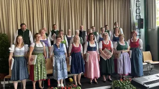 Ein Chor, Traboch, steht auf der Bühne, lächelt und hält Musikbücher. Sie tragen traditionelle Kleidung. Dahinter hängt ein Banner mit einem Musiknotenzeichen über einem Vorhang.