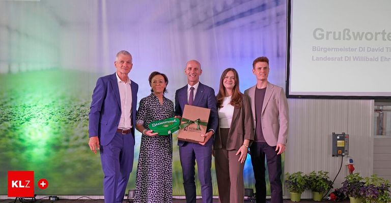 Fünf Personen stehen lächelnd auf einer Bühne und halten eine Geschenkbox, eine Frau hält eine grüne Box. Hinter ihnen zeigt eine Projektionsleinwand ein grünes Feld.