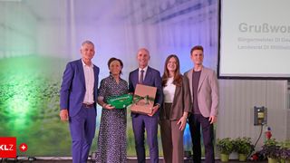 Five people stand on a stage holding a gift box, smiling, with one woman holding a green box. Behind them, a projector screen shows a green field.