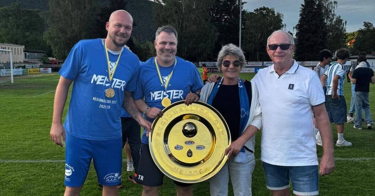 Four people stand on a grassy field, smiling and posing with a gold trophy. Two men wear blue shirts with the word Meister, and one man wears a white shirt. A woman in a black shirt and blue scarf holds the trophy. Trees and a blurry field are behind them.