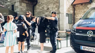 Mehrere Polizeibeamte unterstützen eine Person in Schwarz, möglicherweise einen Demonstranten, in einer Außenumgebung mit einer Steinmauer und geparkten Fahrzeugen.
