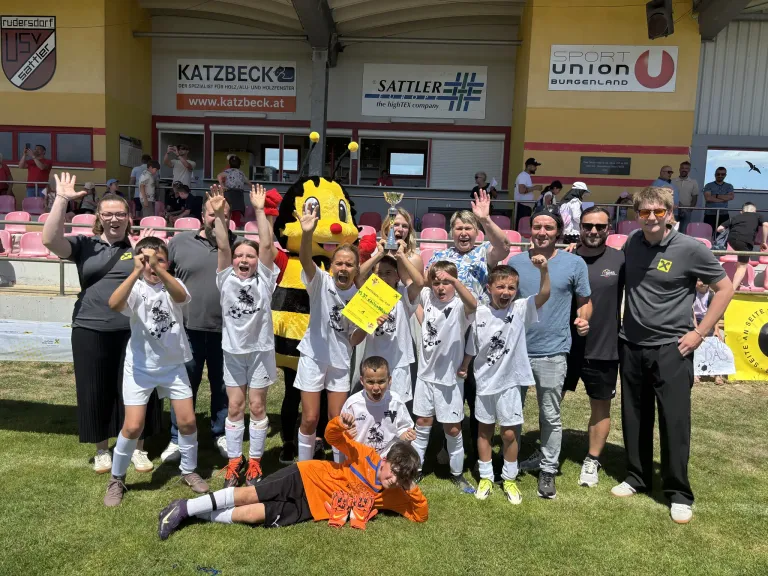 Eine Gruppe von Kindern und Erwachsenen feiert ein Fußballspiel. Sie halten einen Pokal und posieren für ein Foto. Ein Maskottchen, das als Biene verkleidet ist, steht zwischen ihnen.