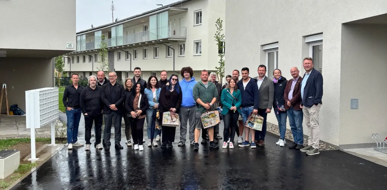 A group of people stand in front of an apartment building in the rain, all holding bags and smiling for a photo.