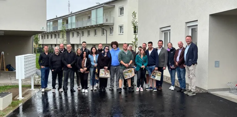 A group of people stand in front of an apartment building in the rain, all holding bags and smiling for a photo.