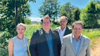 Vier Personen stehen draußen auf einem Feld. Der Mann rechts hat ein breites Lächeln. Der Mann in der Mitte hat die Hände in den Taschen. Die Frau links trägt ein blaues Kleid. Der Hintergrund ist voller Bäume.