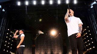 Two men on stage, one with tattoos sings into a microphone while the other holds a microphone. Bright stage lights and a blurred background.