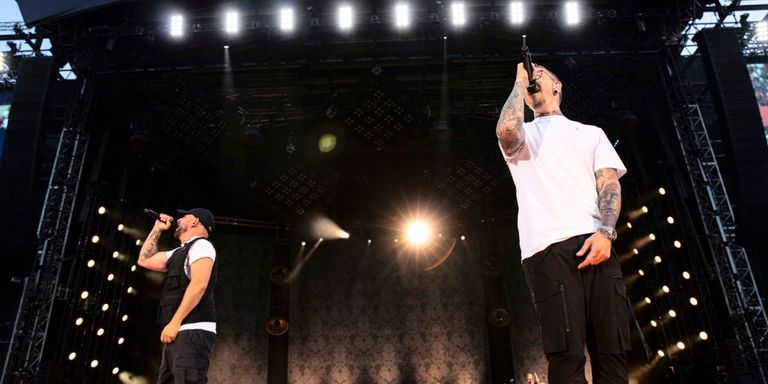 Two men on stage, one with tattoos sings into a microphone while the other holds a microphone. Bright stage lights and a blurred background.