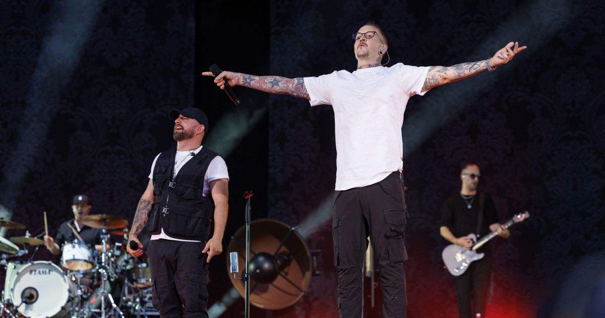 A man with tattoos on his arms sings on stage while another man plays the drums and a guitarist stands behind him.