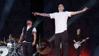 A man with tattoos on his arms sings on stage while another man plays the drums and a guitarist stands behind him.