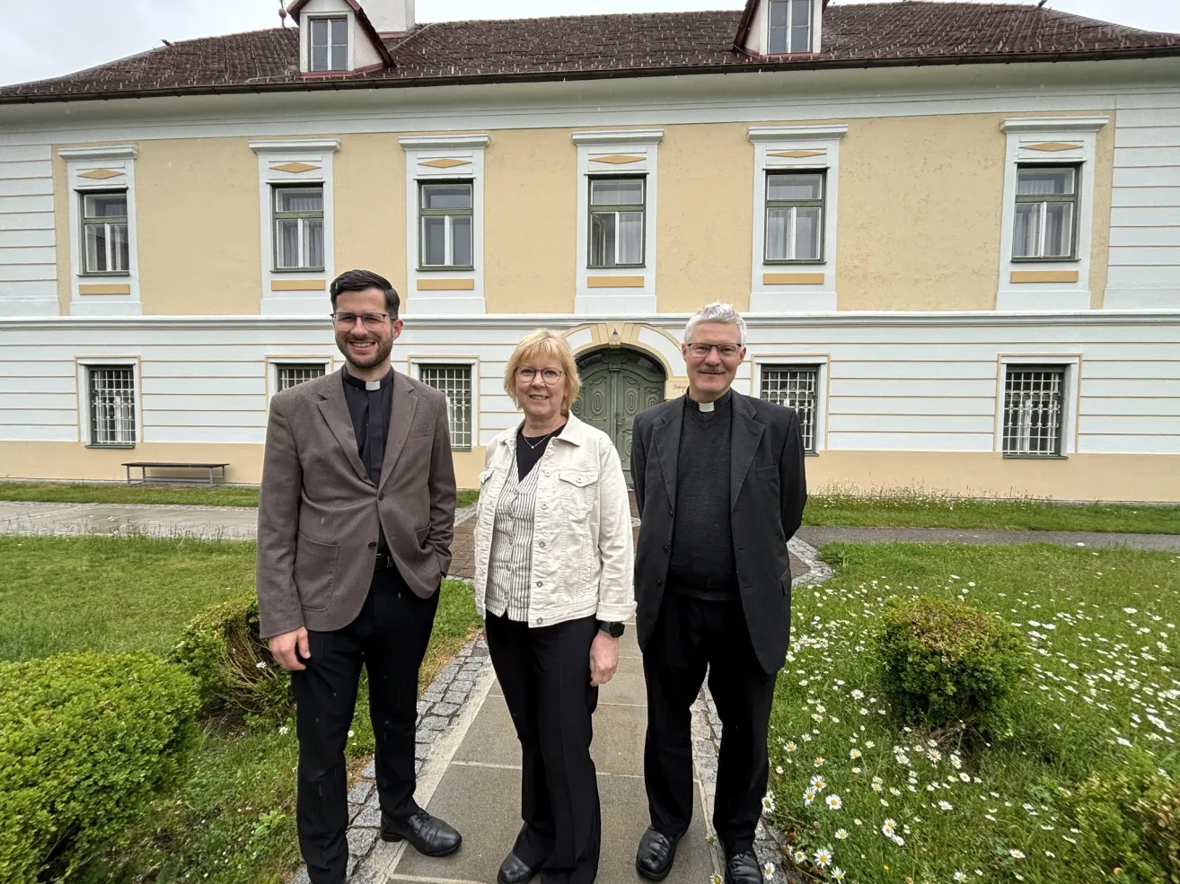 Three people stand in front of a building. Two men wear black clerical collars and suits, while the woman wears a white jacket. They all smile for the camera.