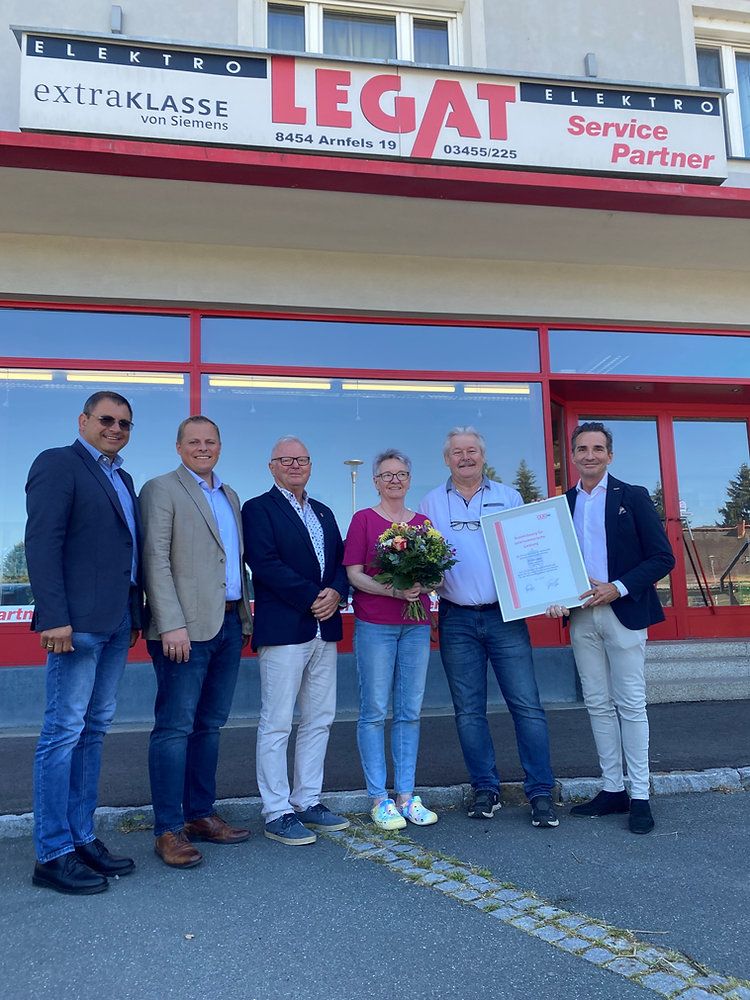 Six people pose for a photo outside a store with a woman holding a bouquet of flowers and a man holding a certificate.