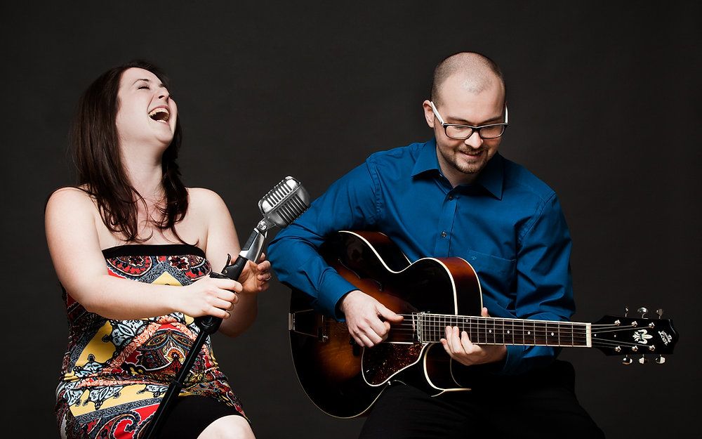 A woman singing into a microphone and a man playing a guitar, both smiling.