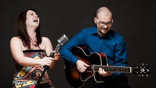 A woman singing into a microphone and a man playing a guitar, both smiling.