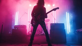 A person with long hair stands on stage, playing a guitar under pink and purple lights.