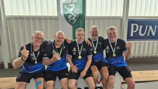 Five men in blue jerseys with medals around their necks sit on a bench, smiling for a photo. Behind them is a banner with a coat of arms.
