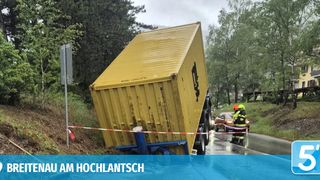 A large yellow shipping container has fallen onto its side in a forested area. Two emergency workers are standing nearby, and a car is parked behind them. The area is marked with barriers.
